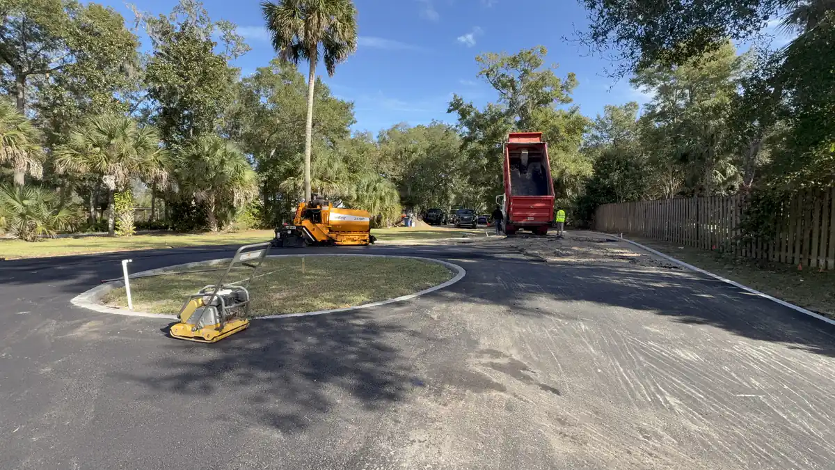 Dump truck raising its bed to feed the paver on a DeLand cul-de-sac, with the crew working around the centre island