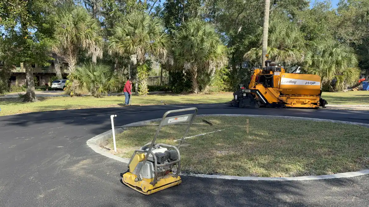 LeeBoy paver laying asphalt around the centre island of a DeLand cul-de-sac beside a walk-behind plate compactor