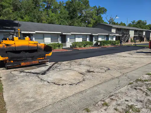 During paving: yellow paver beside the worn gray old surface of a Daytona Beach apartment lot, with fresh black asphalt already laid along the unit fronts