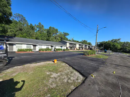 During paving: roller compacting fresh black asphalt along the unit fronts of a Daytona Beach apartment complex beside yellow caution tape marking a closed section