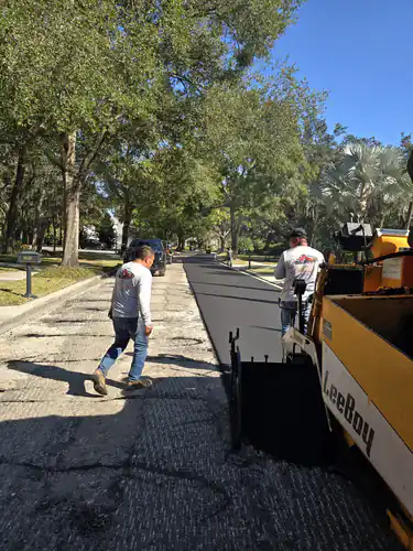 LeeBoy paver laying fresh asphalt under live oaks in Sanford while a crew member walks the raw base ahead of the machine