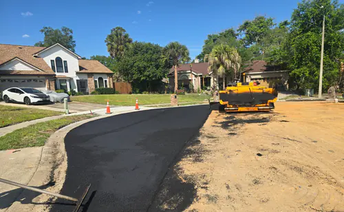 During paving: paver at the bare dirt end of a Maitland cul-de-sac with fresh black asphalt running back toward the street between orange traffic cones