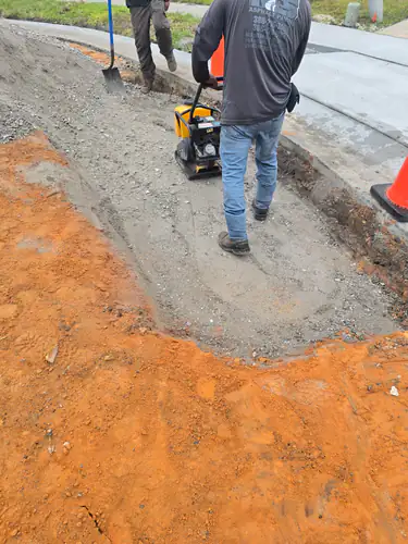 Before paving: crew member walking a plate compactor over crushed stone along a new concrete curb with orange subsoil beside it in Maitland