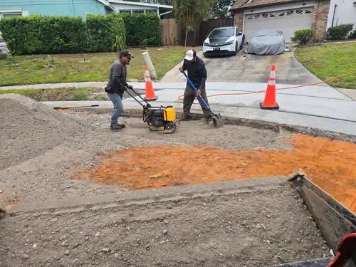 Before paving: two crew members compacting a crushed stone base with a plate compactor beside bare orange subsoil on a Maitland cul-de-sac