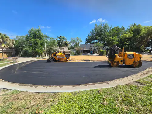 During paving: roller with M&S lettering compacting the new asphalt arc of a Maitland cul-de-sac with the paver and bare ground behind it