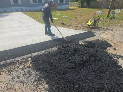 During repair: crew member raking a pile of hot asphalt along the edge of a new concrete driveway in Ormond Beach with a plate compactor on the grass