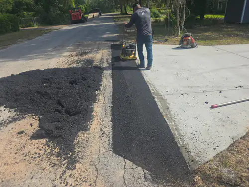 During repair: crew member compacting a strip of fresh asphalt along a new concrete driveway apron in Ormond Beach with a pile of hot mix on the left