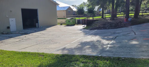 Before: pale cracked driveway beside a metal outbuilding in Ormond Beach with a fenced yard and a dumpster in the background