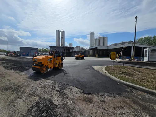 During paving: Cat tandem roller on fresh asphalt in an Orlando industrial yard, with cracked patched old pavement in the foreground and silos behind