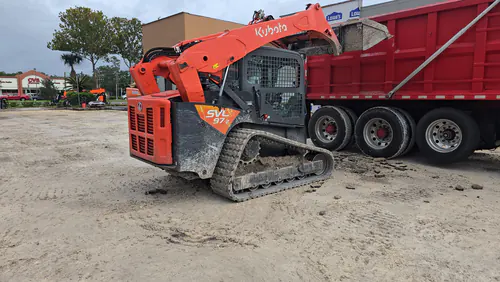 Kubota compact track loader tipping spoil into a red dump truck on a parking lot build at an Orlando shopping complex