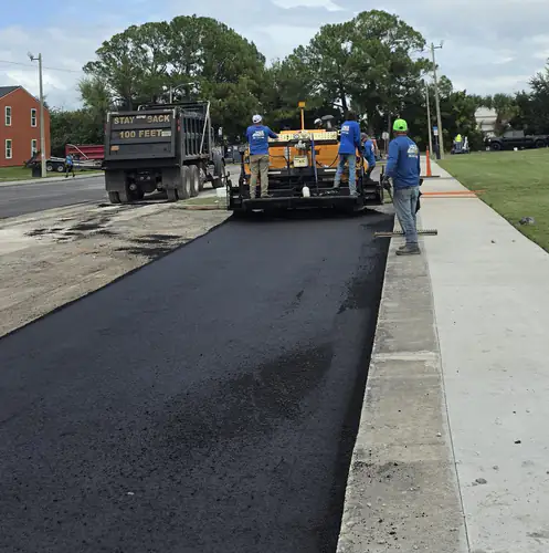 Crew member raking the edge of fresh asphalt against a concrete sidewalk behind the paver at an apartment complex in DeBary, Florida