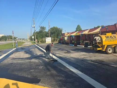 M&S Asphalt Paving crew member spreading sealcoat with a squeegee at the entrance of the Los Dos Compadres parking lot in DeLand, beside the M&S tank trailer