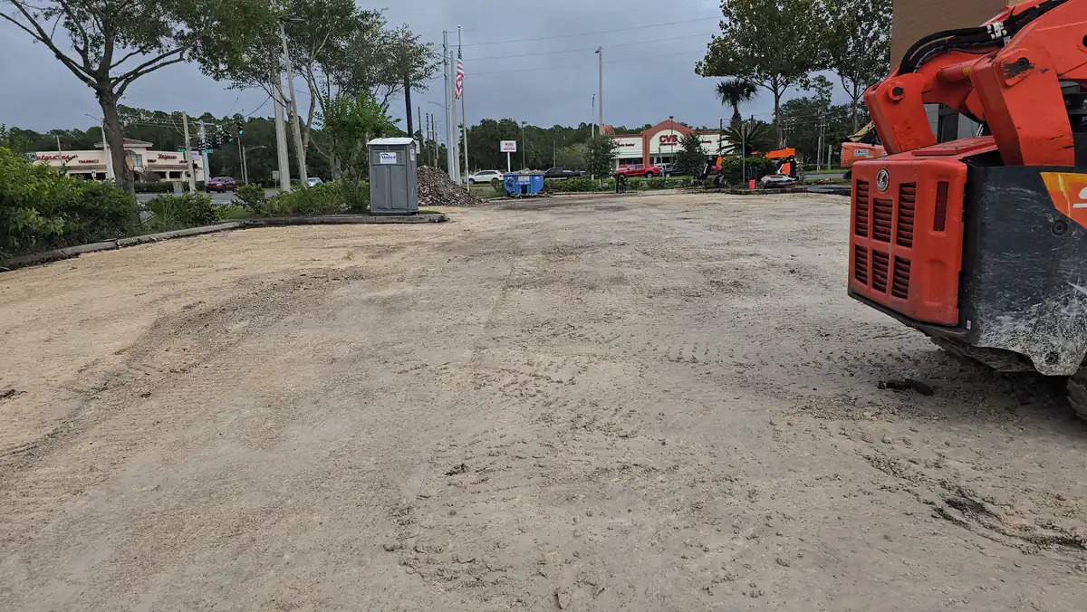 Graded sand subgrade of a new parking lot at an Orlando shopping complex, with a compact track loader at the right and storefronts behind