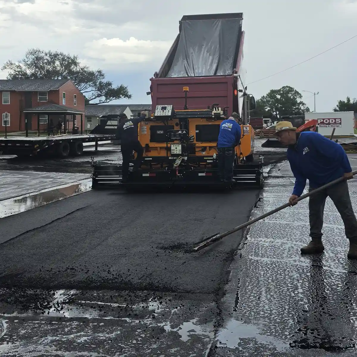 Crew member luting the edge of fresh asphalt on wet pavement behind the paver at an apartment complex parking lot in DeBary, Florida