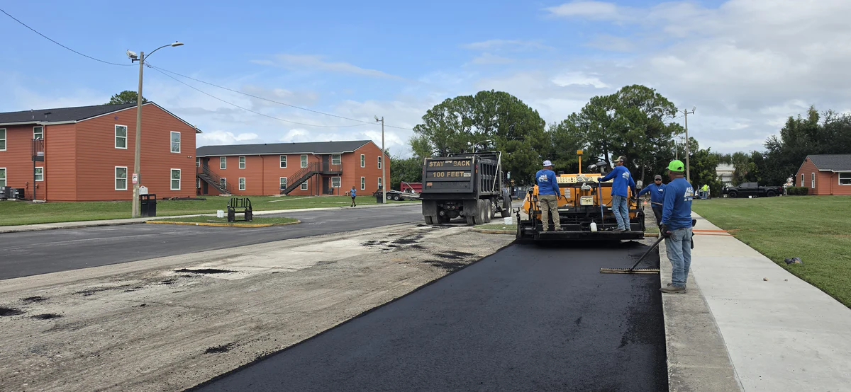 Long strip of fresh asphalt along a concrete sidewalk at a DeBary apartment complex, with the paver, dump truck, and crew at the far end and an unpaved section to the left