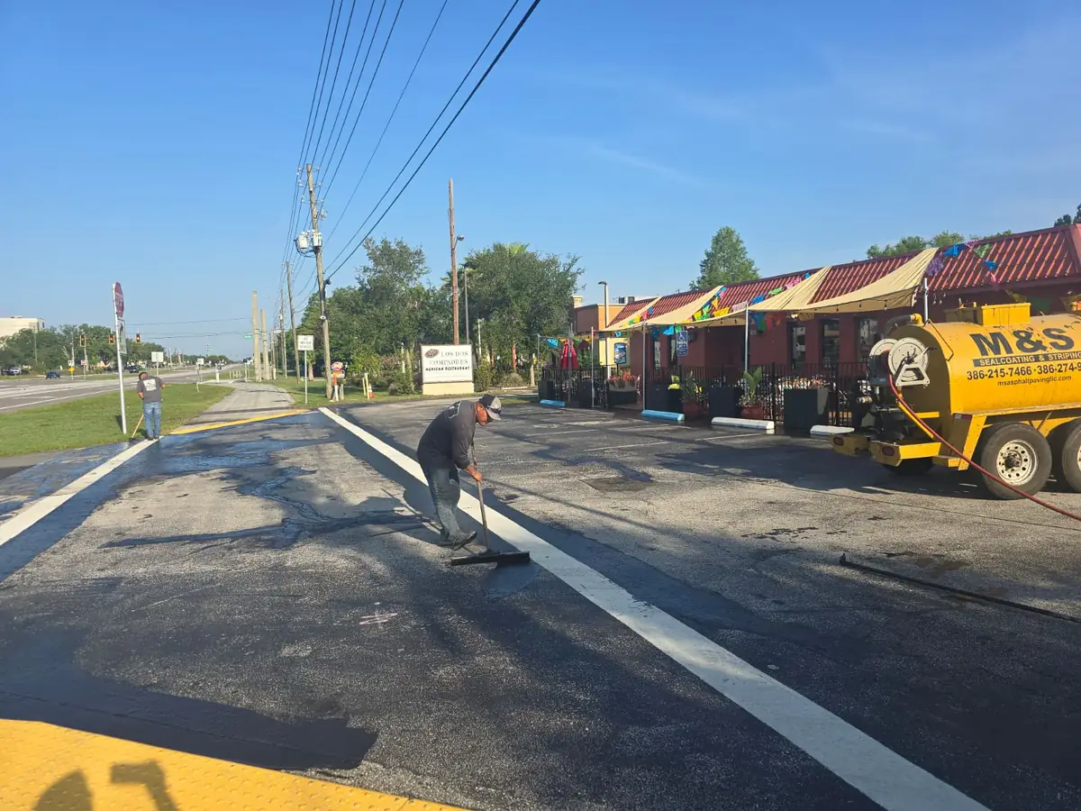 Crew member spreading sealcoat with a squeegee at the Los Dos Compadres parking lot entrance in DeLand, Florida, next to the M&S tank trailer