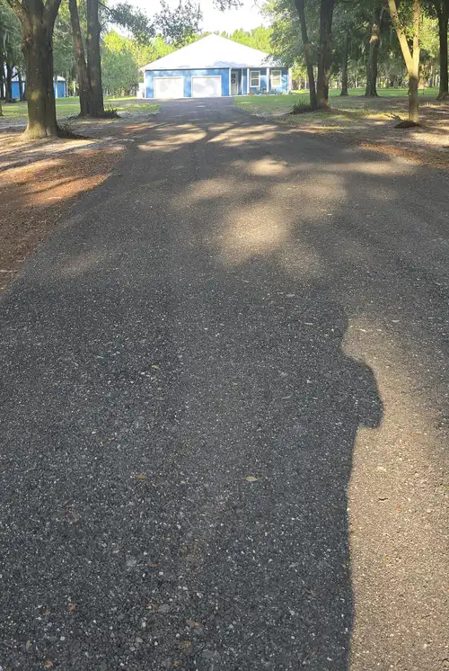 Finished recycled asphalt millings driveway leading to a blue house through oak trees