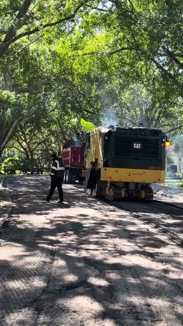 Cold planer grinding the old asphalt off a shaded DeLand street with a dump truck alongside it